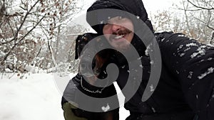 An owner with his dog is hugging and making selfie in a winter wood among falling snow in slowmo