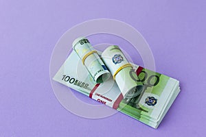 A pack of 100 euro banknotes in a bank package and two rolls under an elastic band on it. lilac background