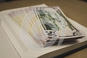 A pack of dollars and a notebook open with a pencil on a wooden background