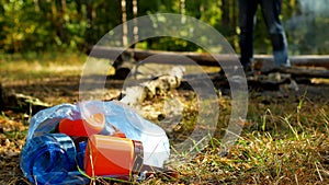 A package with plastic garbage lies on a glade, a fire burns, people walk in the background, camping and nature