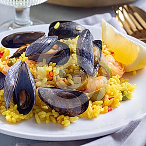 Paella with mussels and shrimps, on white plate, with lemon and white wine, square, closeup