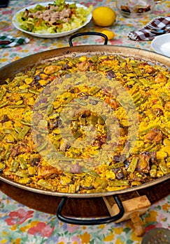 Paellera with a freshly cooked authentic Valencian paella, to serve on a table with the dishes set, salad, glasses and the photo