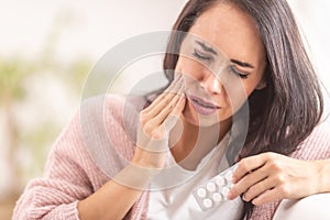 Pain in tooth while brunette holds her cheek with one hand, and painkilling pills in the other hand