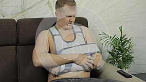 Painful, bored man with a broken arm wearing arm brace sitting on a sofa