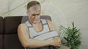 Painful, bored man with a broken arm wearing arm brace sitting on a sofa