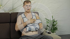 Painful, bored man with a broken arm wearing arm brace sitting on a sofa