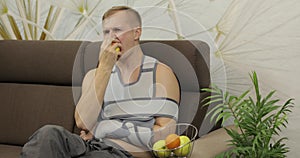 Painful man with a broken arm wearing arm brace sitting on a sofa eating apple