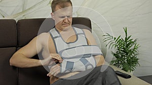 Painful man with a broken arm wearing arm brace sitting on a sofa watching TV