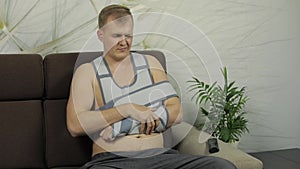 Painful man with a broken arm wearing arm brace sitting on a sofa watching TV