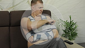 Painful man with a broken arm wearing arm brace sitting on a sofa watching TV