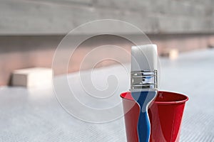 Paint brush and a bucket with paint next to a building exterior wall. Painting house plinth outside. Home renovation. Nobody