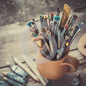 Paintbrushes in a jug from potters clay, wooden palette and paints.