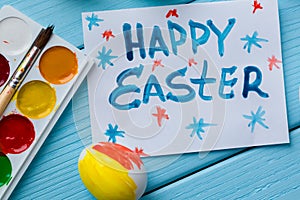 Painted Easter egg, paints, paintbrush and note with title written by paint `Happy Easter` on the blue colored wooden background