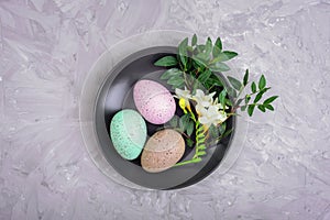 Painted Easter eggs and freesia white flower on black plate on gray textured background. Holiday table setting. Top view