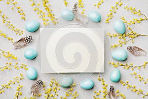 Painted Easter eggs, mimosa flower and paper card on vintage stone background top view in flat lay style. Empty space for text.