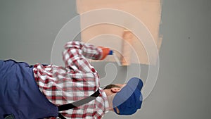 Painter in blue dungarees, cap and gloves painting a wall with paint roller in new house