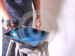 The painter dips his brush in paint bath of blue paint. Cuvette for rollers on a stepladder.