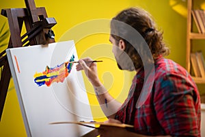 A painter is looking directly into the camera and smiling as he is holding up his art palette and sitting in a chair in