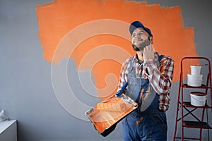 Painter man holding a paint roller and tray having doubts and with confuse face expression standing near painting wall