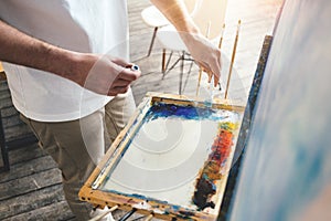 Painter mixes oil paint colors on the palette in workshop. Men artist in light studio