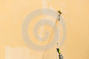 Painter paints building exterior wall with a roller. Roller with long stick manually painting building with yellow paint