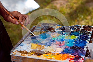 Painter`s skilful hand mixes oil paints with a brush on wooden palette, stained with a fresh stained colorful mess