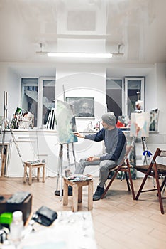 Painter sits on a chair in a cozy studio and works on a painting, drawing oil paintings. Artist works in a home painting studio.