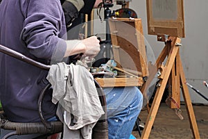 Painter sits near easel with open box of paint colors