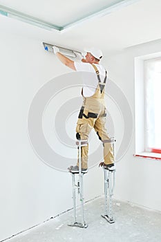 Painter in stilts with putty knife. Plasterer smoothing ceiling surface at home renewal