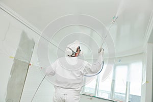 Painter worker with airless painting sprayer covering ceiling surface into white photo