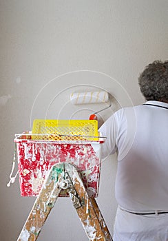 Painter working in a room. The colorman is painting a wall using a paint roller, paint tray, paint scuttle and a ladder