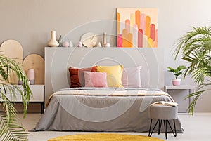Painting on grey headboard of bed with colorful cushions in bedroom interior with stool. Real photo