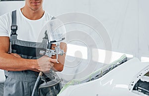 With painting gun. Caucasian automobile repairman in uniform works in garage