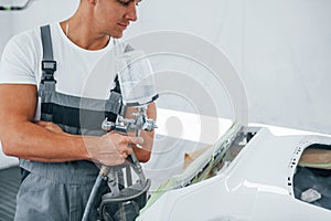 With painting gun. Caucasian automobile repairman in uniform works in garage