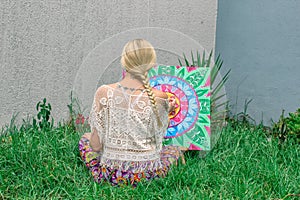 Painting outdoors, a young woman blonde draws a mandala on the nature sitting in the grass