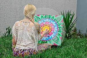 Painting outdoors, a young woman blonde draws a mandala on the nature sitting in the grass