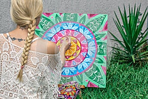 Painting outdoors, a young woman blonde draws a mandala on the nature sitting in the grass