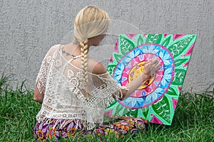 Painting outdoors, a young woman blonde draws a mandala on the nature sitting in the grass