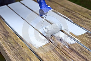 Painting a weathered wooden garden table with a paint roller and white wood protection coat, restorating old furniture for the