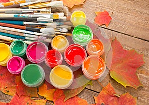 Paints, brushes, autumn leaves on wooden background.