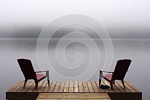Pair of Adirondack chairs on wooden deck on lake in fog with copy space