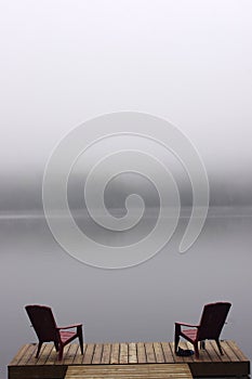 Pair of Adirondack chairs on wooden deck on lake in fog with copy space