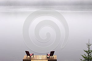 pair of Adirondack chairs on wooden deck on lake in fog with copy space