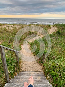 A pair of bare feet, crossed ankles pointing down a set of weathered steps to a strip of sandy path which leads to the ocean.