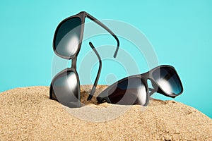 Pair of black sunglasses vertically sticking out of sand on blue.