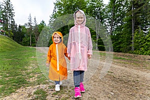Pair of cute little caucasian sibling kids in bright waterproof raincoats walking under rain in scenic moody dirt forest
