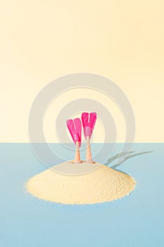 A pair of diving fins pokes out of the sand on blue and yellow pastel background. Creative idea of summer swimming recreation, or