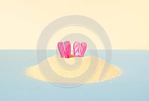 A pair of diving pink fins pokes out of the sand on blue and yellow pastel background. Creative idea of summer swimming undersea