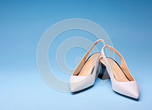 A pair of elegant white pointed-toe female shoes with an open back and black flare heels on a gradient blue background. Stylish