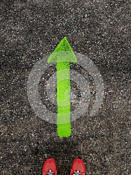 A pair of feet wearing red sport shoes standing on tarmac road with green arrow print sign pointing toward